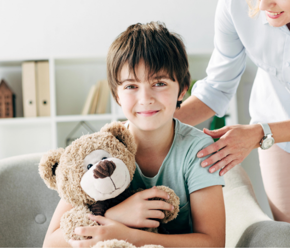 Child hugging a teddy bear with a supportive adult by his side
